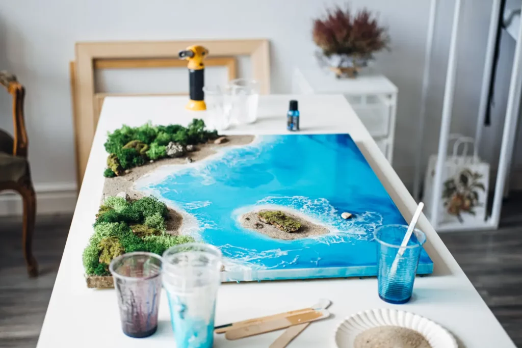 Blue resin art canvas, miniature island, and shoreline scene, craft supplies on table.