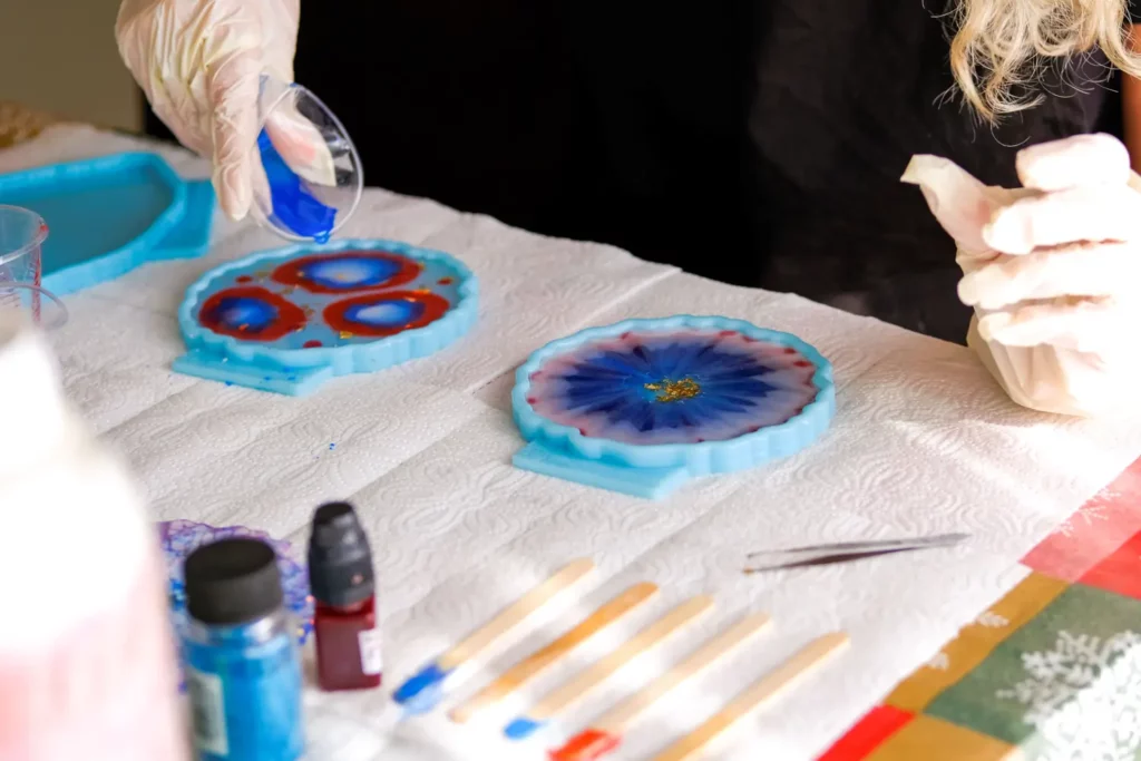 Person in gloves pouring resin into silicone molds, creating colorful resin art coasters.  Blue, red, and gold colors.
